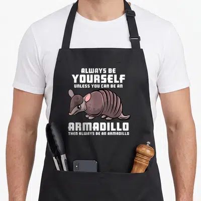 Alway armadillo apron - Cartoon armadillo in purple-brown with white text quote about being yourself — shown on a black cotton canvas apron against a white background