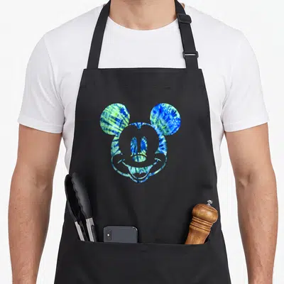 Funny tie dye apron - Mickey Mouse head silhouette filled with blue green and yellow tie-dye pattern on black fabric — shown on a black cotton canvas apron against a white background