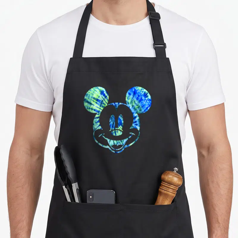 Funny tie dye apron - Mickey Mouse head silhouette filled with blue green and yellow tie-dye pattern on black fabric — shown on a black cotton canvas apron against a white background
