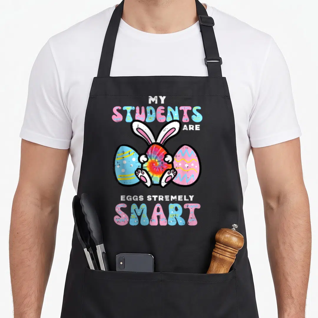 Eggstremely teacher apron - Bunny peeking from behind three eggs with My Students Are Eggs Tremely Smart text — shown on a black cotton canvas apron against a white background