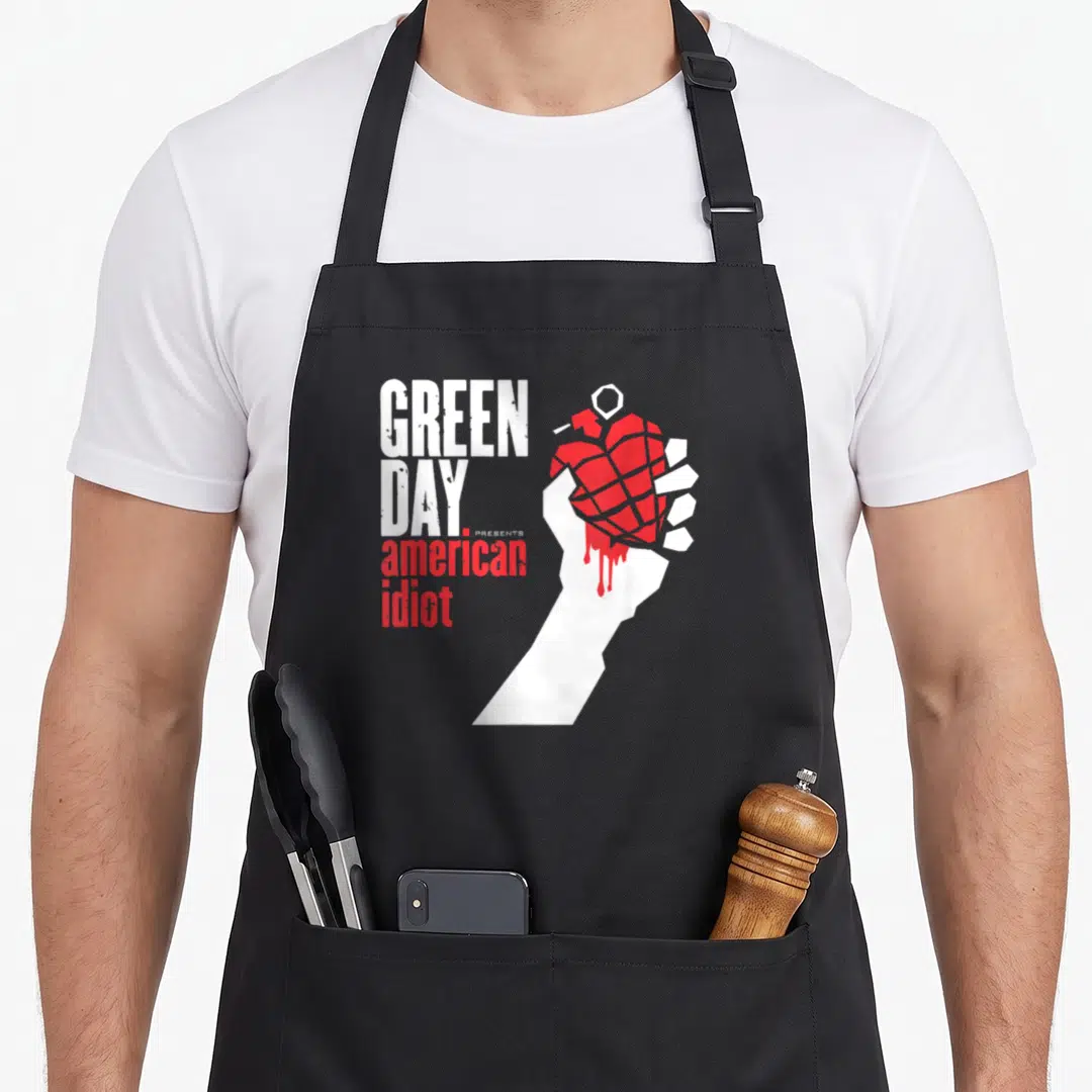 Idiot band apron - White fist holding red dripping heart grenade next to stacked Green Day American Idiot text — shown on a black cotton canvas apron against a white background