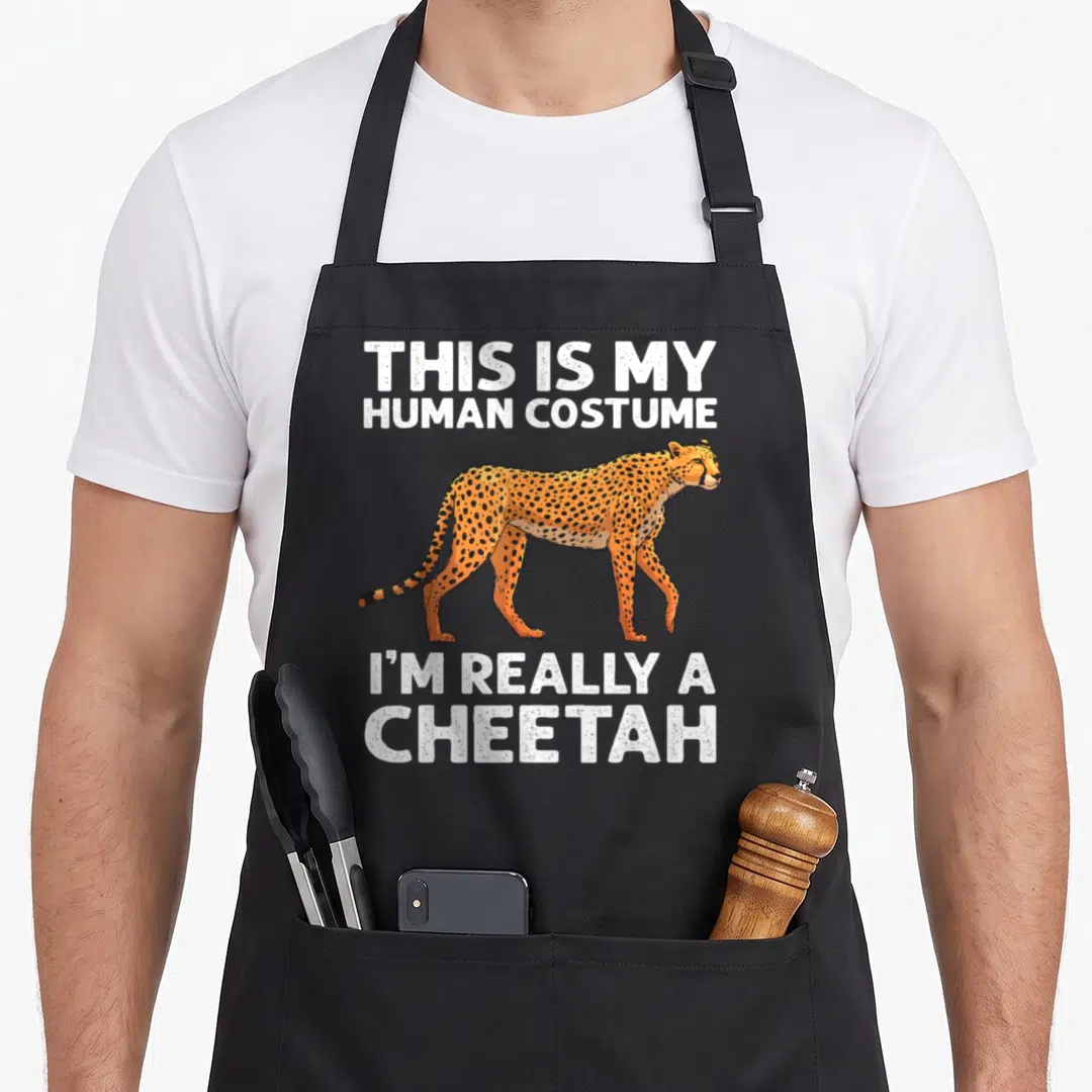 Wildlife cheetah apron - Orange cheetah walking between white text blocks reading human costume joke — shown on a black cotton canvas apron against a white background