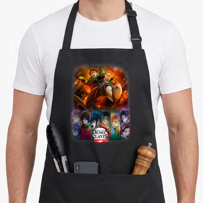 Infinity anime apron - Tanjiro Kamado leaping over Demon Slayer Infinity Castle logo and vertical character portraits — shown on a black cotton canvas apron against a white background