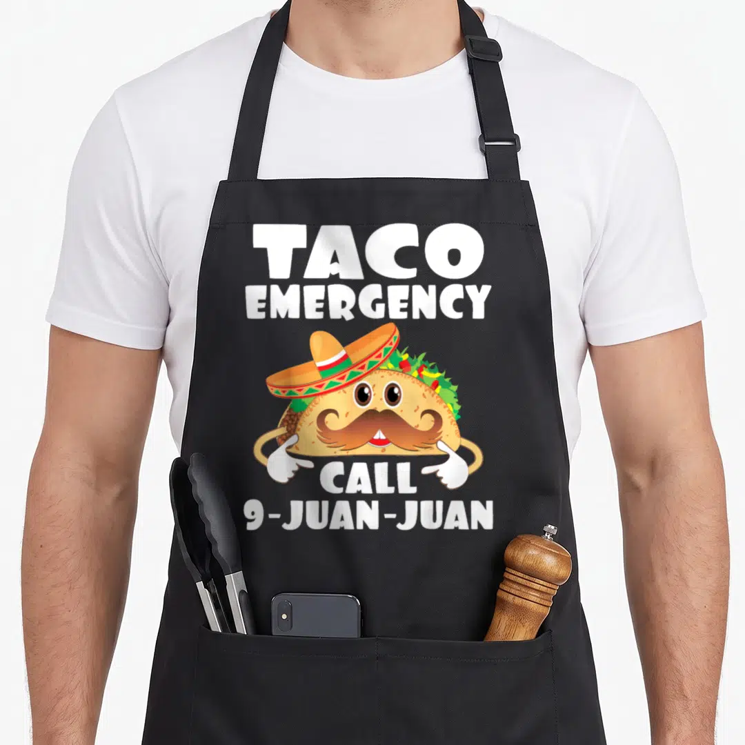 Call taco apron - Cartoon taco with sombrero and mustache pointing to TACO EMERGENCY CALL 9-JUAN-JUAN text — shown on a black cotton canvas apron against a white background