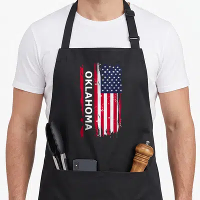 Funny oklahoma boy apron - Vertical OKLAHOMA text overlaid on a distressed red white and blue USA flag graphic — shown on a black cotton canvas apron against a white background