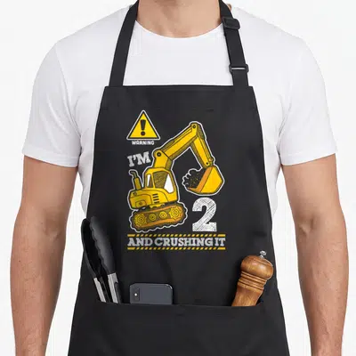 Digger truck apron - Cartoon excavator with WARNING I'M 2 AND CRUSHING IT text in yellow and white — shown on a black cotton canvas apron against a white background