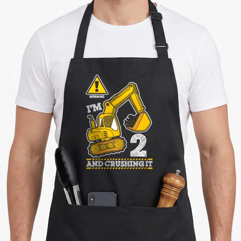 Digger truck apron - Cartoon excavator with WARNING I'M 2 AND CRUSHING IT text in yellow and white — shown on a black cotton canvas apron against a white background