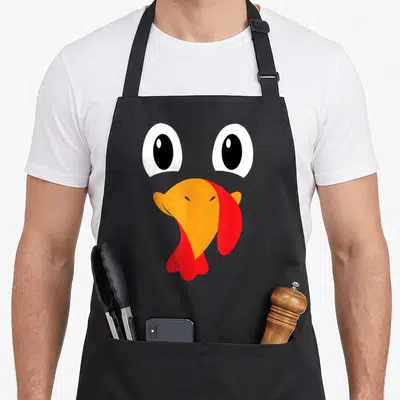 Halloween turkey apron - Cartoon turkey face with two white eyes, orange beak, and red wattle — shown on a black cotton canvas apron against a white background