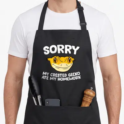 Lizard pet apron - Cartoon crested gecko head with sorry my crested gecko ate my homework text — shown on a black cotton canvas apron against a white background