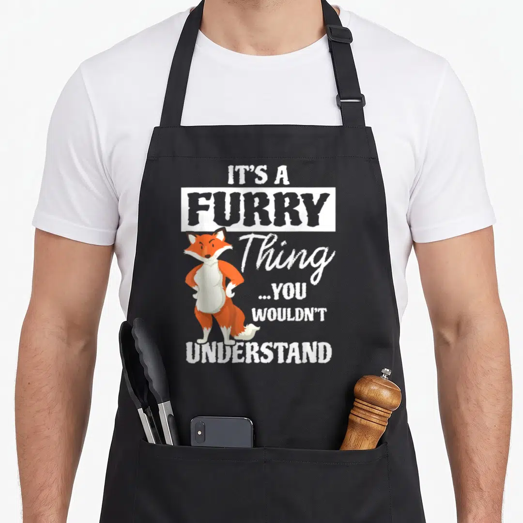 Furry animal apron - Orange cartoon fox standing next to white stacked text reading IT'S A FURRY Thing — shown on a black cotton canvas apron against a white background