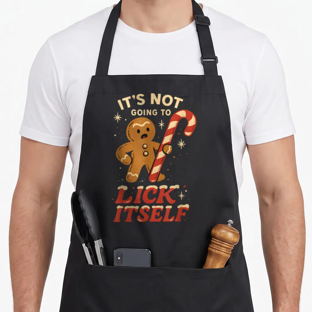 Candy gingerbread apron - Cartoon gingerbread man holding red candy cane with IT'S NOT GOING TO LICK ITSELF text — shown on a black cotton canvas apron against a white background