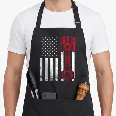 Car american flag apron - Red piston graphic overlaid on white stripes and grey stars in a distressed style — shown on a black cotton canvas apron against a white background