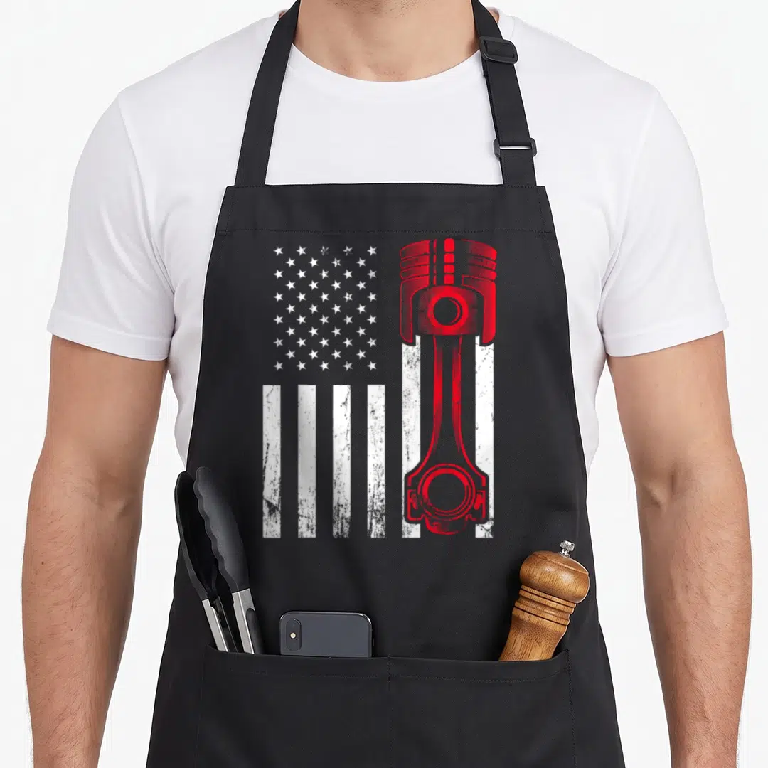 Car american flag apron - Red piston graphic overlaid on white stripes and grey stars in a distressed style — shown on a black cotton canvas apron against a white background