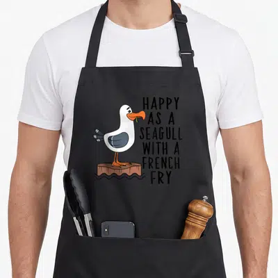 Clothe french apron - Cartoon seagull holding yellow fry on brown post next to stacked black text reading happy as — shown on a black cotton canvas apron against a white background
