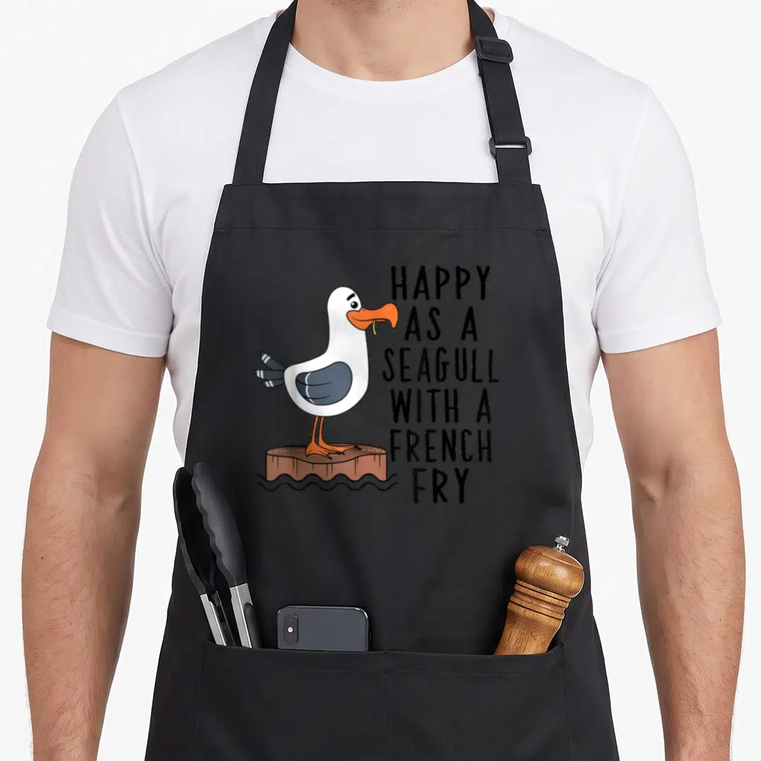 Clothe french apron - Cartoon seagull holding yellow fry on brown post next to stacked black text reading happy as — shown on a black cotton canvas apron against a white background