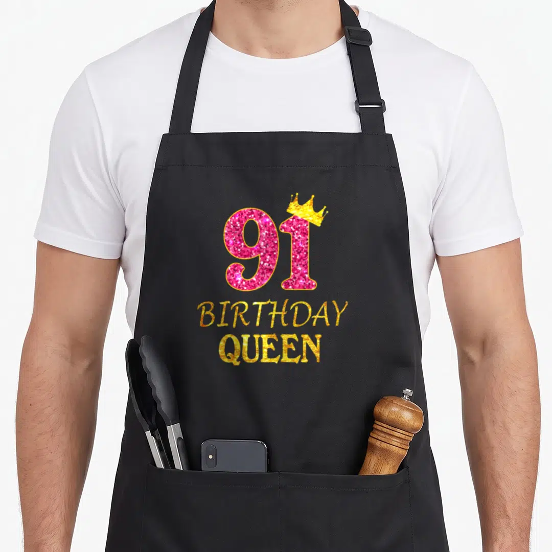 91st queen apron - Pink 91 with gold crown and gold BIRTHDAY QUEEN text — shown on a black cotton canvas apron against a white background