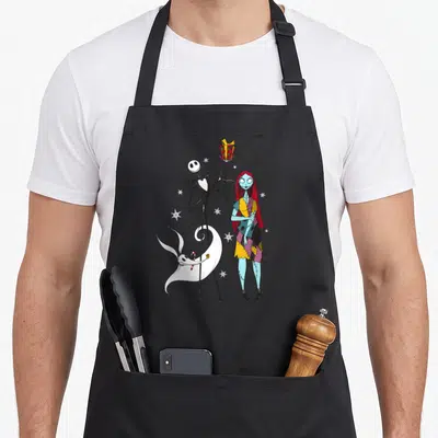 Jack zero lover apron - Jack holds red gift, Sally stands right, Zero floats with candy cane, white snowflakes scattered — shown on a black cotton canvas apron against a white background