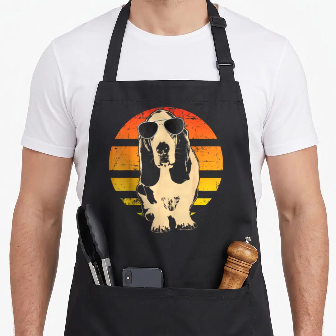 Basset dog apron - Basset hound in sunglasses before orange yellow striped sun graphic — shown on a black cotton canvas apron against a white background