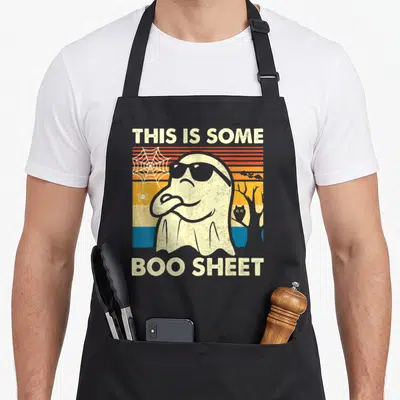 Sheet halloween apron - Retro ghost with sunglasses and 'THIS IS SOME, BOO SHEET' text over striped background — shown on a black cotton canvas apron against a white background