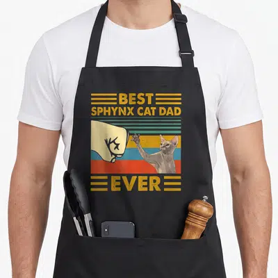 Sphynx sunset cat apron - Retro fist bump between human hand and sphynx cat with best sphynx cat dad ever text — shown on a black cotton canvas apron against a white background