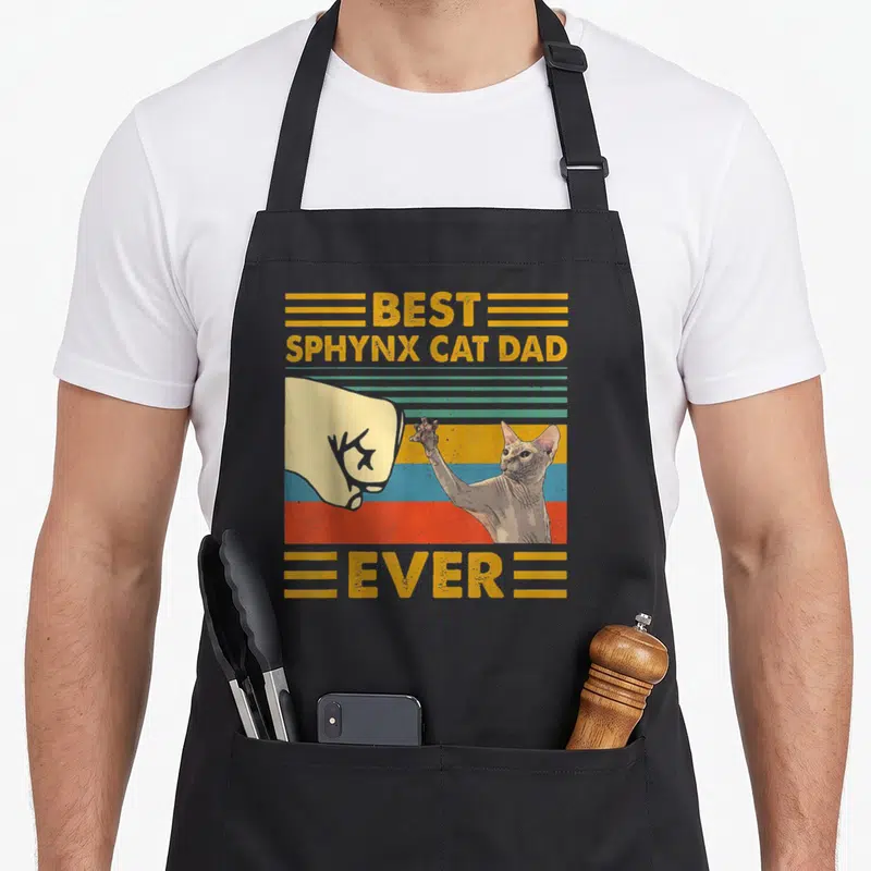 Sphynx sunset cat apron - Retro fist bump between human hand and sphynx cat with best sphynx cat dad ever text — shown on a black cotton canvas apron against a white background