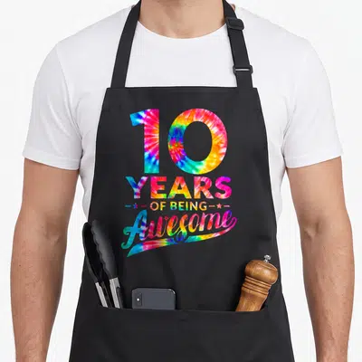10th birthday tie dye apron - Stacked 10 YEARS OF BEING Awesome text in tie dye with two stars — shown on a black cotton canvas apron against a white background