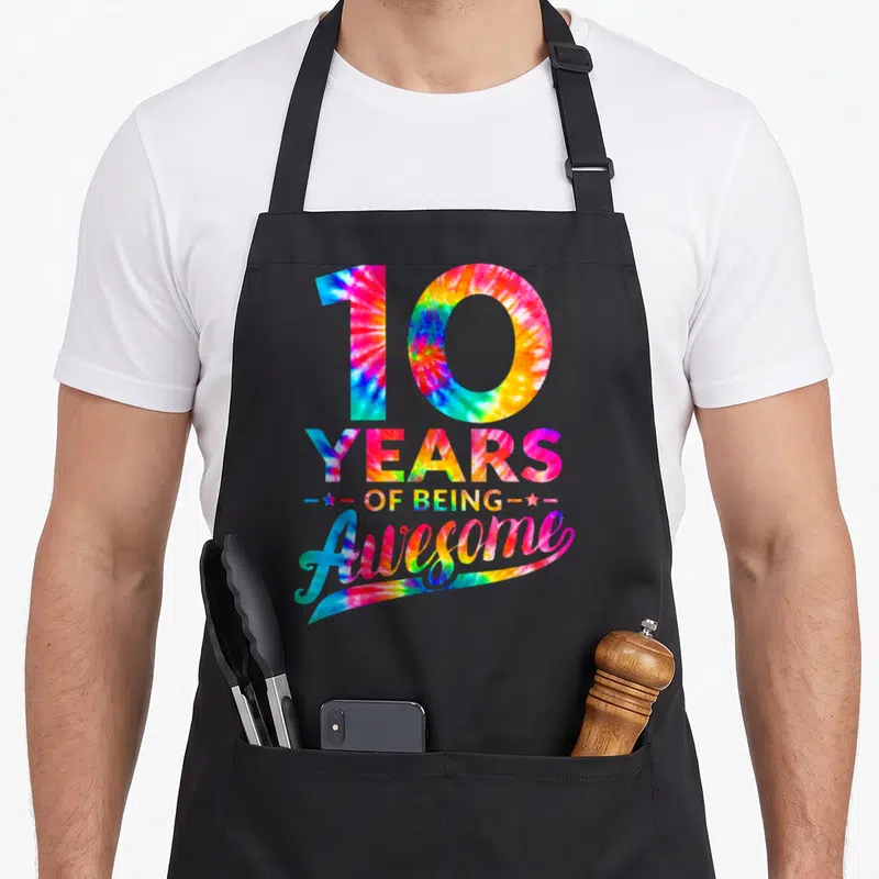 10th birthday tie dye apron - Stacked 10 YEARS OF BEING Awesome text in tie dye with two stars — shown on a black cotton canvas apron against a white background