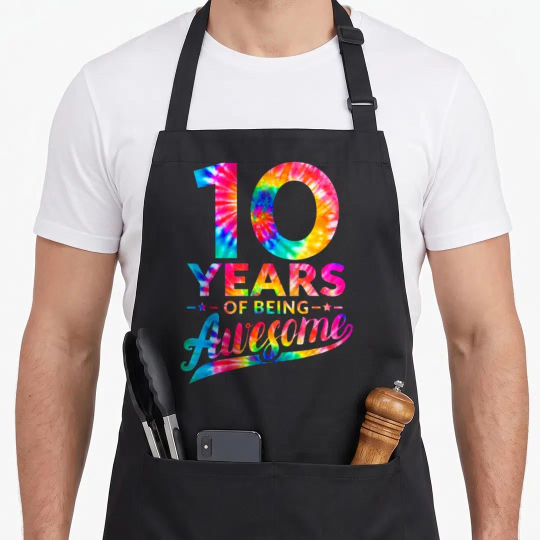 10th birthday tie dye apron - Stacked 10 YEARS OF BEING Awesome text in tie dye with two stars — shown on a black cotton canvas apron against a white background
