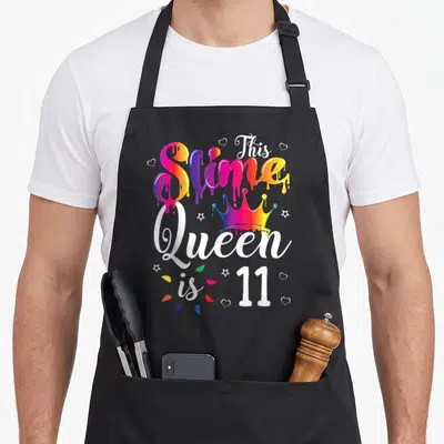 Slime queen of baking apron - Rainbow slime text and crown reading This Slime Queen is 11 — shown on a black cotton canvas apron against a white background