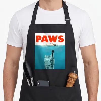 Meow kitten apron - Red PAWS text above underwater cat reaching up toward swimmer in blue water — shown on a black cotton canvas apron against a white background