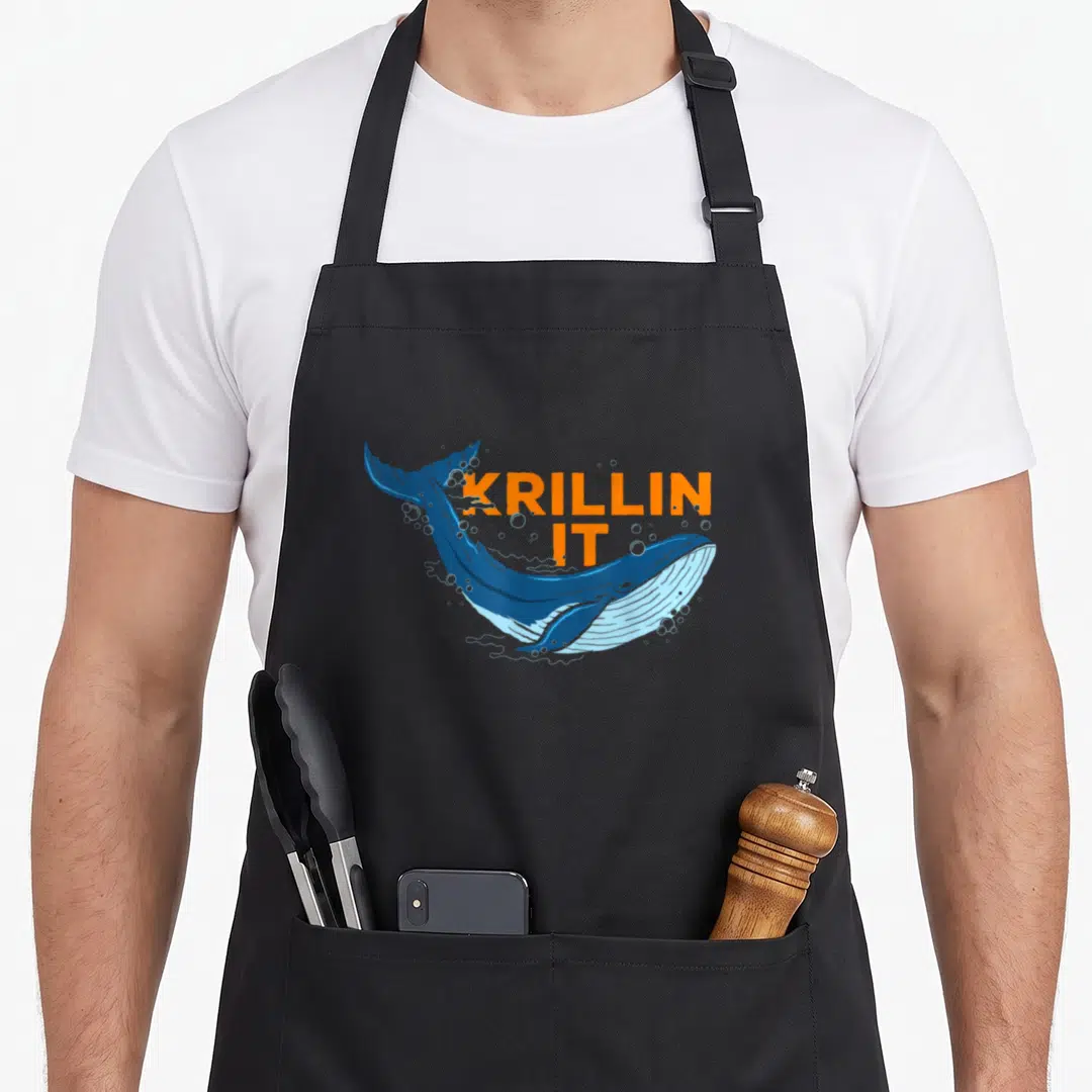 Humpback animal apron - Cartoon blue whale leaping with orange KRILLIN IT text overlay — shown on a black cotton canvas apron against a white background