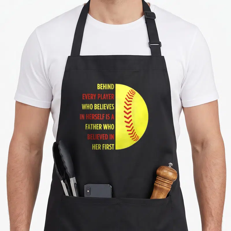 Behind father daughter aprons - Stacked yellow and red text next to a yellow half-softball with red stitching — shown on a black cotton canvas apron against a white background