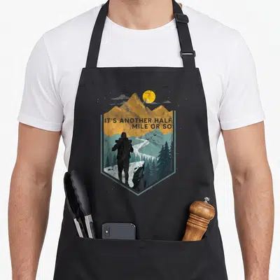 Another apron or apron - Retro hiker silhouette facing mountains with text IT'S ANOTHER HALF MILE OR SO in yellow — shown on a black cotton canvas apron against a white background