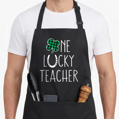 Prek st patrick day aprons - One Lucky Teacher text with plaid shamrock and white horseshoe typography — shown on a black cotton canvas apron against a white background