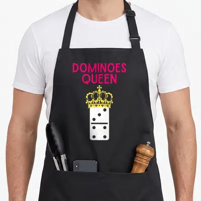 Player queen apron - Pink DOMINOES QUEEN text above yellow crown on white domino tile — shown on a black cotton canvas apron against a white background
