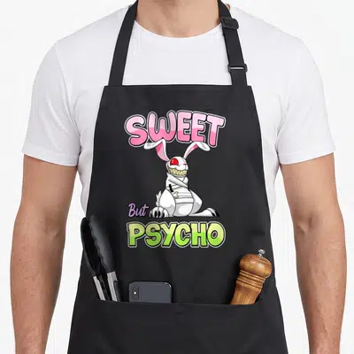Sweet gothic apron - Cartoon rabbit with sharp teeth and SWEET But PSYCHO text in pink and lime green — shown on a black cotton canvas apron against a white background