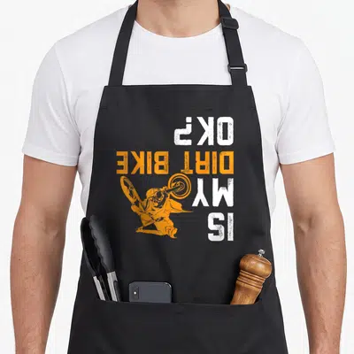 Motocross bike apron - Orange rider silhouette overlapping vertical white and orange text reading Is My Dirt Bike OK — shown on a black cotton canvas apron against a white background