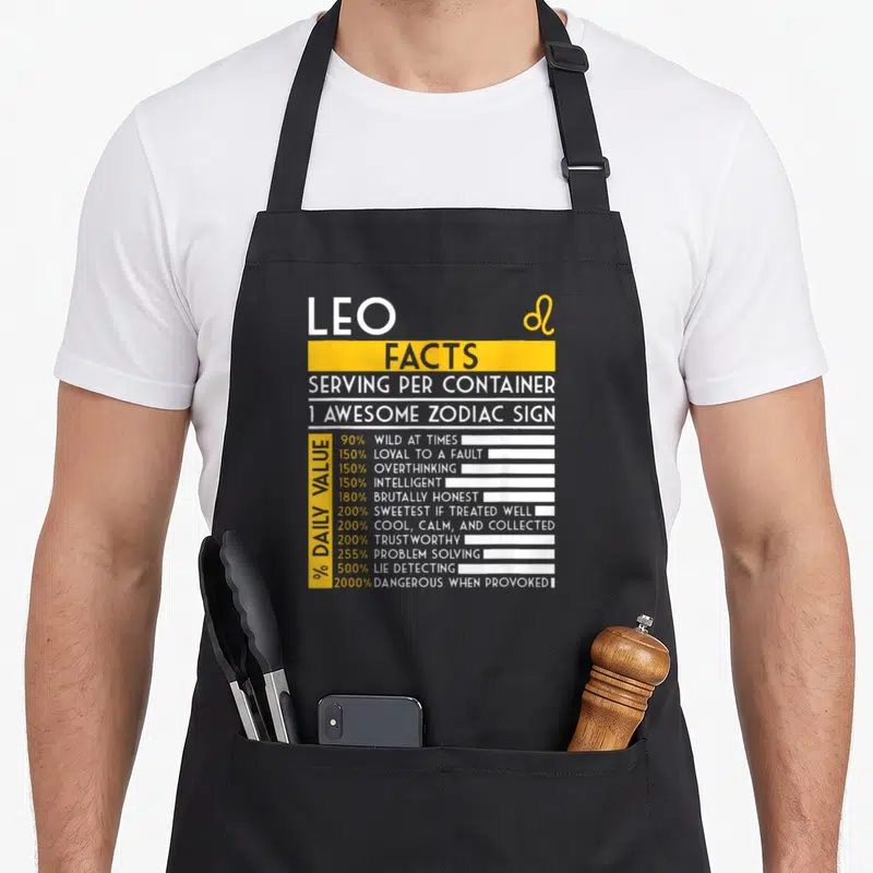 Leo fact zodiac aprons - Nutrition label style graphic listing Leo traits like 500% lie detecting and 2000% dangerous — shown on a black cotton canvas apron against a white background
