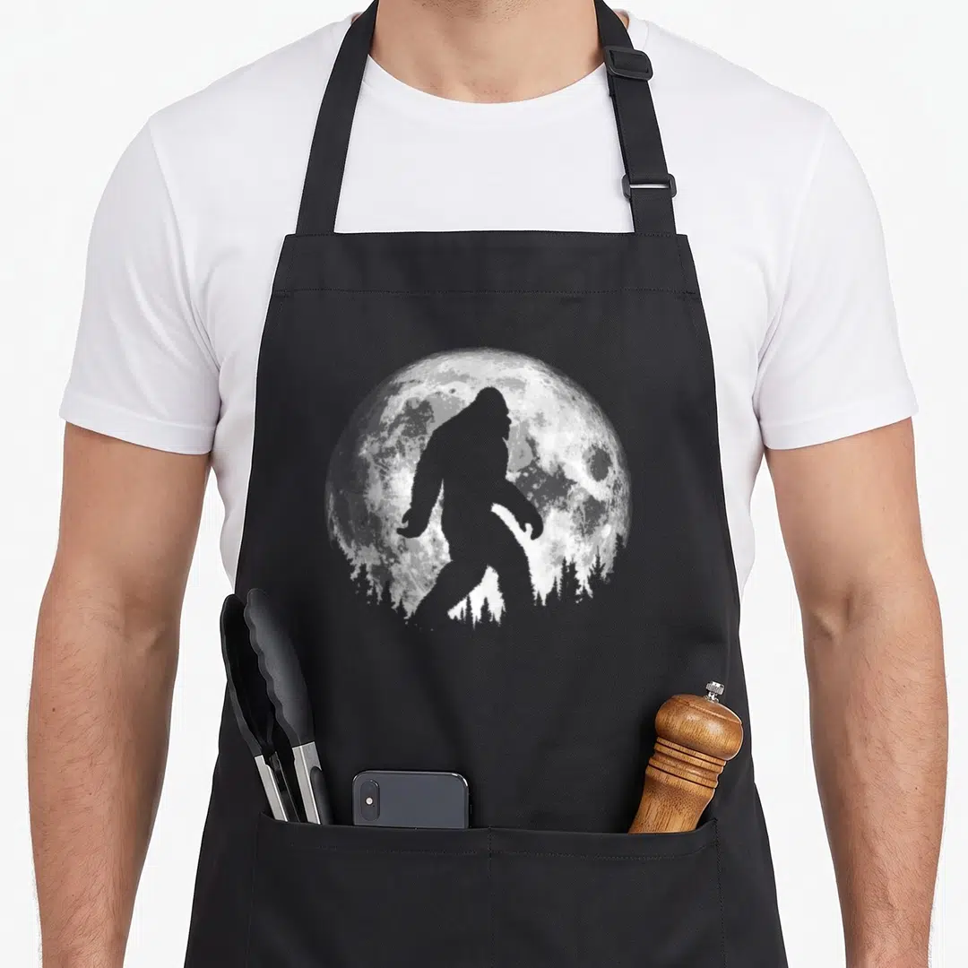 Bigfoot tree apron - Grey bigfoot silhouette walking in front of a white textured moon with pine trees at the bottom — shown on a black cotton canvas apron against a white background