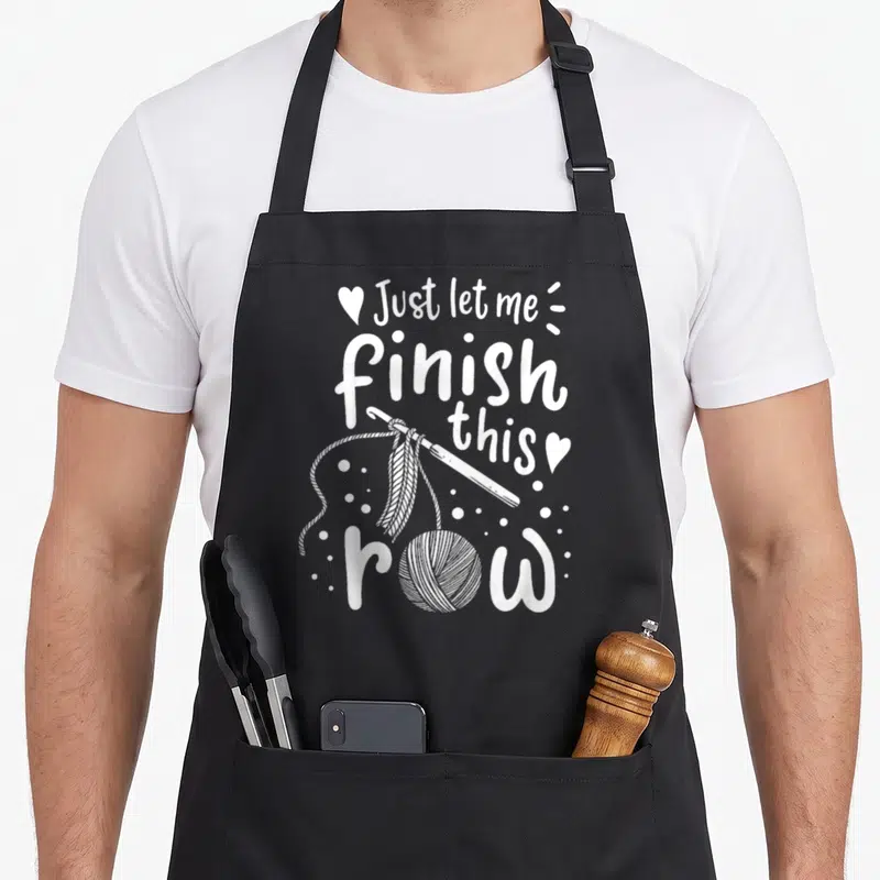 Funny crocheting knitting apron - Just let me finish this row with crochet hook and yarn ball in white typography — shown on a black cotton canvas apron against a white background