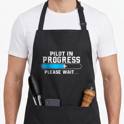 Progress aircraft apron - Distressed white Pilot In Progress text above blue loading bar with white airplane and Please — shown on a black cotton canvas apron against a white background