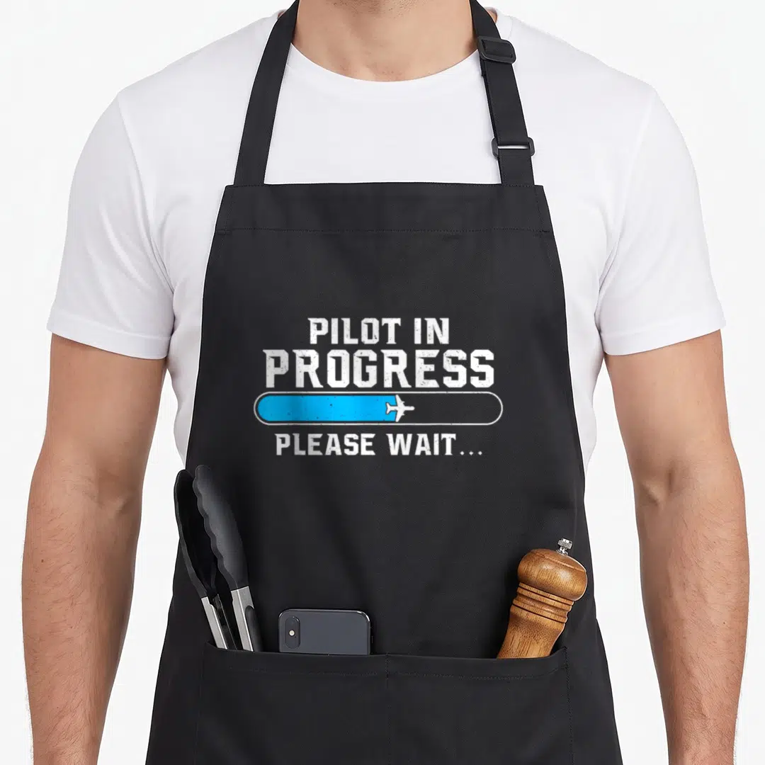 Progress aircraft apron - Distressed white Pilot In Progress text above blue loading bar with white airplane and Please — shown on a black cotton canvas apron against a white background