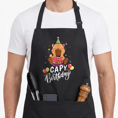 Funny capy birthday apron - Cartoon capybara holding pink cake with white CAPY Birthday text and balloons — shown on a black cotton canvas apron against a white background