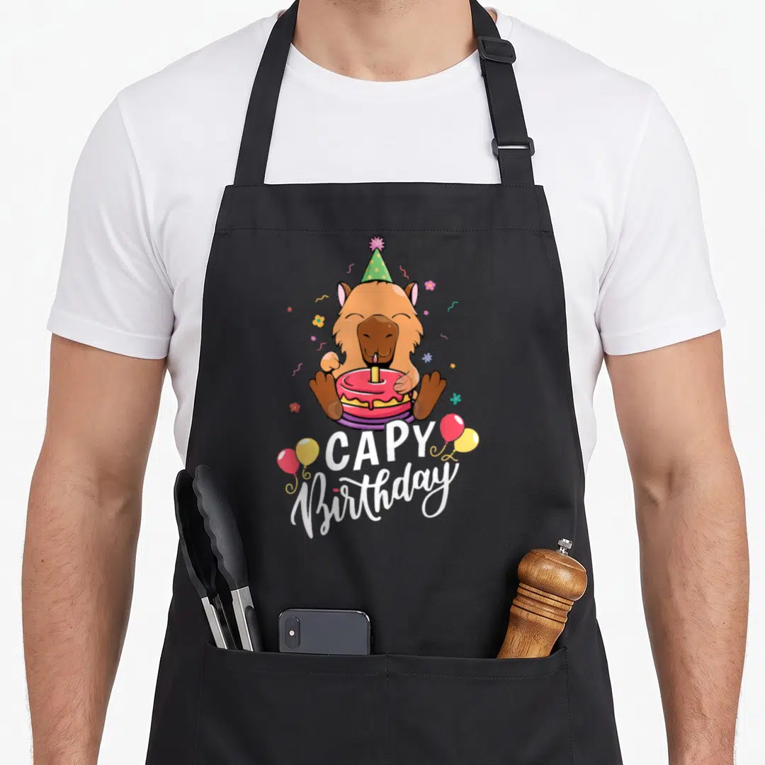 Funny capy birthday apron - Cartoon capybara holding pink cake with white CAPY Birthday text and balloons — shown on a black cotton canvas apron against a white background