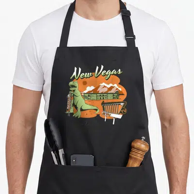 Dino video game apron - Green dinosaur holding motel sign with New Vegas text and No Vac indicator — shown on a black cotton canvas apron against a white background