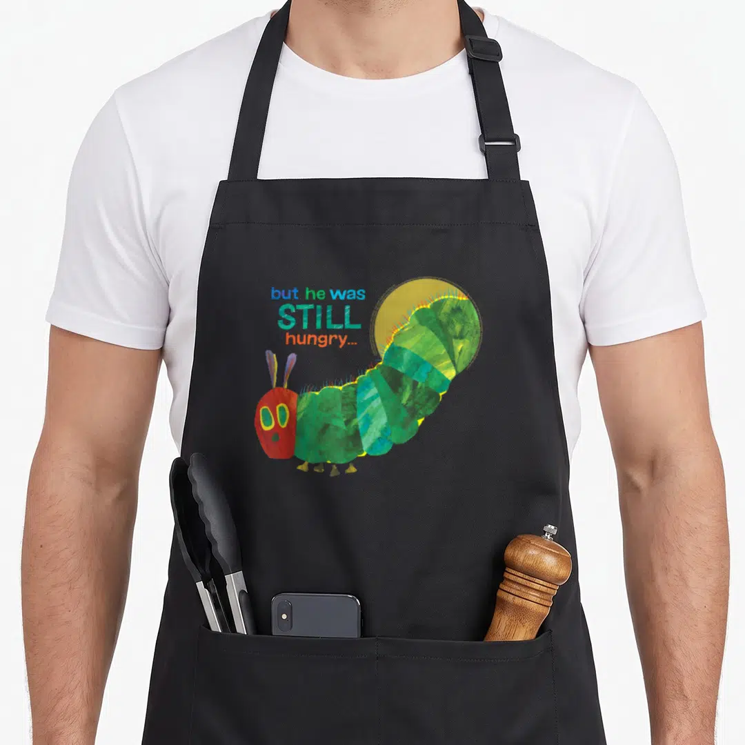 Carle book apron - Green caterpillar with red head and text but he was STILL hungry in watercolor art — shown on a black cotton canvas apron against a white background