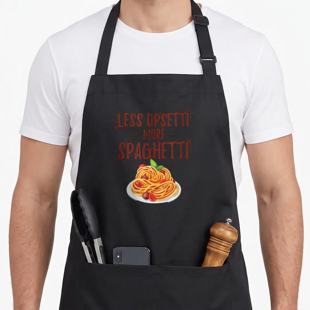Less pasta apron - LESS UPSETTI MORE SPAGHETTI text above cartoon spaghetti plate with tomatoes and basil — shown on a black cotton canvas apron against a white background