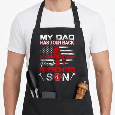 Proud son firefighter aprons - My Dad Has Your Back text with distressed flag and firefighter silhouette flanked by two — shown on a black cotton canvas apron against a white background