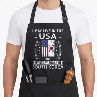South story of the apron - Distressed white text with USA and South Korea flags flanked by wheat ears and stars — shown on a black cotton canvas apron against a white background