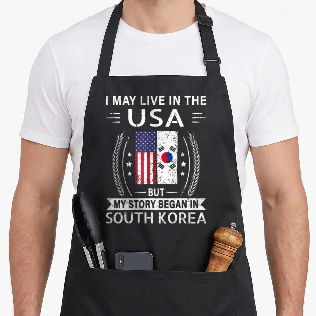 South story of the apron - Distressed white text with USA and South Korea flags flanked by wheat ears and stars — shown on a black cotton canvas apron against a white background